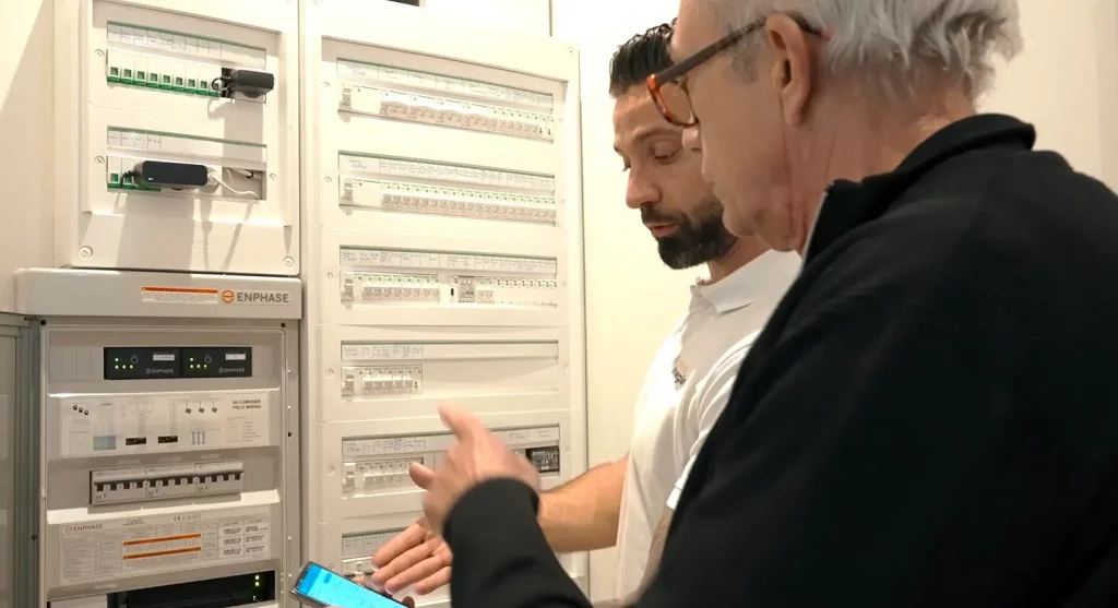 Un expert solaire montre l'application téléphonique de suivi à un client. Ils sont devant un tableau électrique