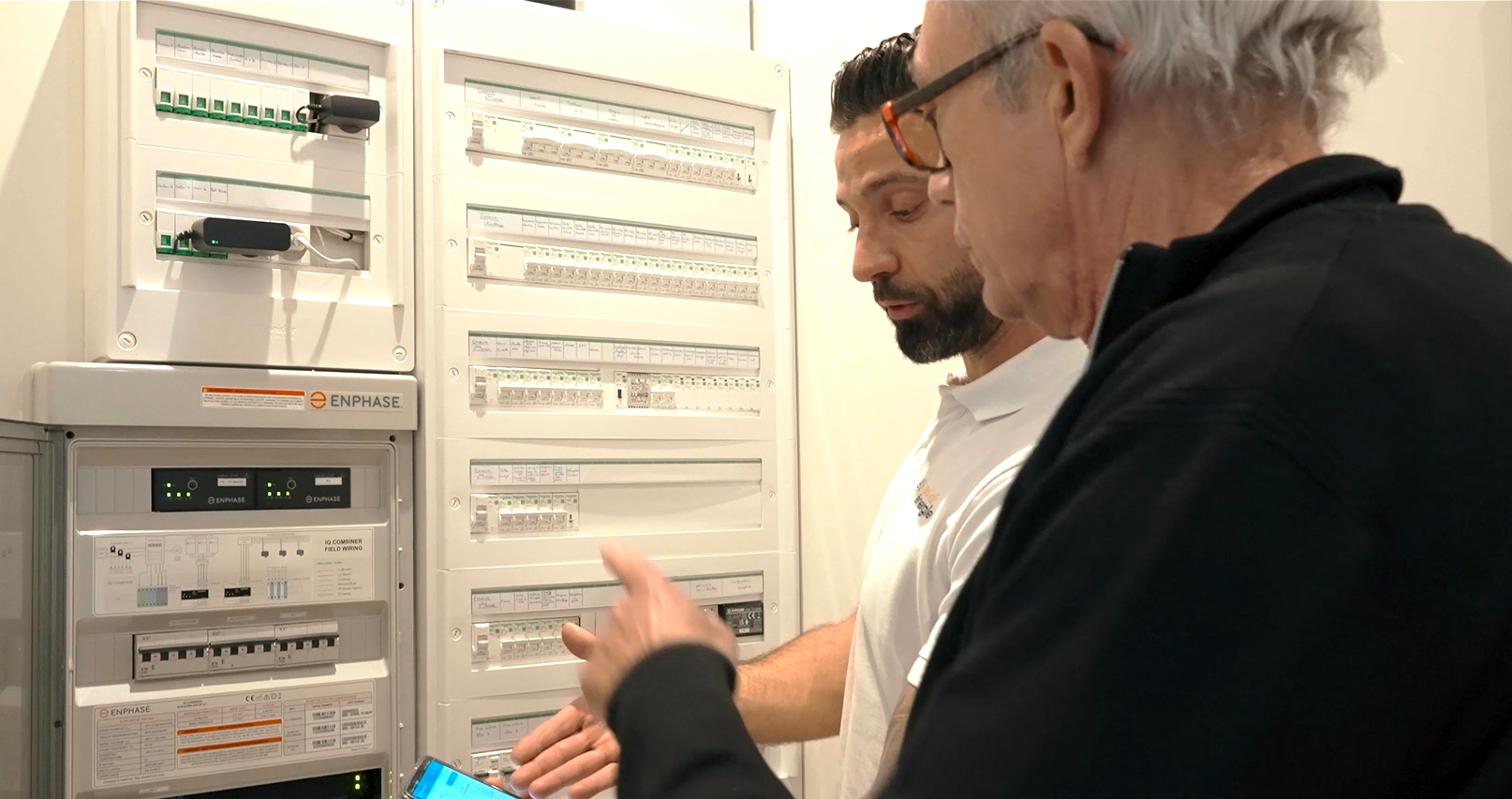 Un expert solaire montre l'application téléphonique de suivi à un client. Ils sont devant un tableau électrique